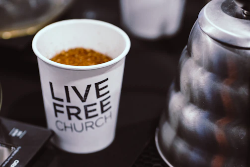A coffee mug with the words 'terrible meeting coffee club' in gold lettering, sitting on a worn church basement table next to a folding chair.