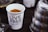 A white cup filled with a brown granular substance, possibly coffee grounds, is prominently displayed. The cup has text printed on it reading 'LIVE FREE CHURCH'. Next to the cup, there is a part of a metallic object, possibly a kettle, with a shiny, reflective surface. The background appears to be a dark surface with blurred elements.