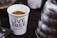 A white cup filled with a brown granular substance, possibly coffee grounds, is prominently displayed. The cup has text printed on it reading 'LIVE FREE CHURCH'. Next to the cup, there is a part of a metallic object, possibly a kettle, with a shiny, reflective surface. The background appears to be a dark surface with blurred elements.