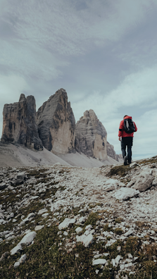 A lone hiker wearing a red jacket and carrying a backpack is walking along a rocky path towards towering, rugged mountain peaks under an overcast sky. The landscape is dotted with rocks and sparse vegetation, creating a sense of isolation and adventure.