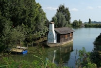 A wooden houseboat with a thatched roof is moored on a serene river, surrounded by lush green trees. The water reflects the vibrant foliage and the clear blue sky above. A nearby wooden dock is visible, partially covered by plants.