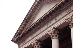 An architectural detail of a classical building featuring ornate Corinthian columns and a triangular pediment. The inscription 'FIDES ET PATRIA' is prominently displayed across the frieze.
