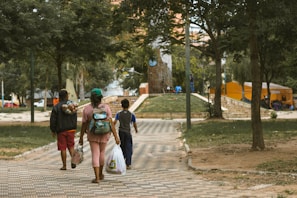 A group of three people, two adults and a child, walking along a paved pathway in a park. The adults are carrying bags, and one has a bundle of clothes over their shoulder. The park is lush with trees and has a few tents set up in the background. The area is well-maintained with green grass and large trees providing shade.