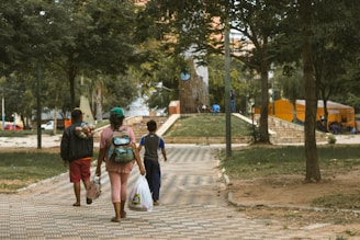 A group of three people, two adults and a child, walking along a paved pathway in a park. The adults are carrying bags, and one has a bundle of clothes over their shoulder. The park is lush with trees and has a few tents set up in the background. The area is well-maintained with green grass and large trees providing shade.