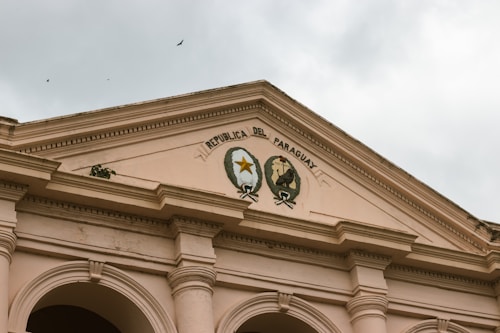 A neoclassical architectural structure with decorative arches and columns is topped by an ornate pediment. The inscription 'Republica del Paraguay' is present along with emblems that include a star and a motto encircled by wreaths. Birds are visible in the overcast sky above.