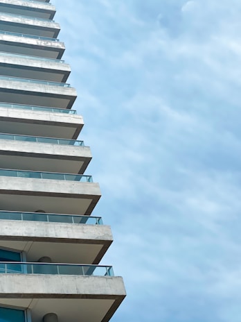 A modern high-rise building with multiple balconies extends upward against a backdrop of a clear blue sky. Each balcony is lined with glass railings, adding to the sleek design of the structure.