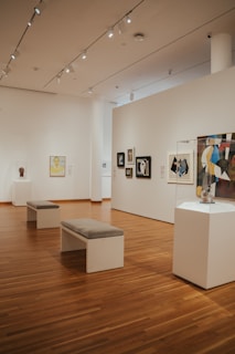 A spacious art gallery interior with wooden floors and several artworks displayed on the walls and pedestals. The ceiling is equipped with bright spotlights. There are two benches in the center for visitors to sit and observe the art pieces.
