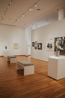 A spacious art gallery interior with wooden floors and several artworks displayed on the walls and pedestals. The ceiling is equipped with bright spotlights. There are two benches in the center for visitors to sit and observe the art pieces.