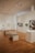 A spacious art gallery interior with wooden floors and several artworks displayed on the walls and pedestals. The ceiling is equipped with bright spotlights. There are two benches in the center for visitors to sit and observe the art pieces.