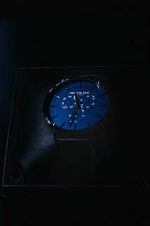 A blue watch is displayed in dim lighting. The watch face includes a chronograph feature with multiple dials and white markings. The brand name is visible at the top of the watch face.
