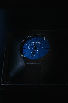 A blue watch is displayed in dim lighting. The watch face includes a chronograph feature with multiple dials and white markings. The brand name is visible at the top of the watch face.