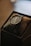 A black wristwatch with a leather strap is resting on a soft cushion inside a box. The watch face has multiple dials and thin silver hands, and the surroundings are softly lit, giving a warm, elegant appearance.