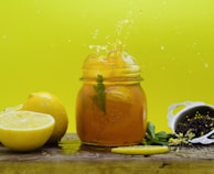 A refreshing scene featuring a mason jar filled with iced tea, garnished with mint leaves and lemon slices, on a wooden surface. A whole lemon and a lemon half are placed beside the jar, adding a vibrant yellow hue to the composition. An overturned metal tea strainer rests nearby, spilling loose black tea leaves. The background is a bright yellow, enhancing the freshness and energy of the scene.