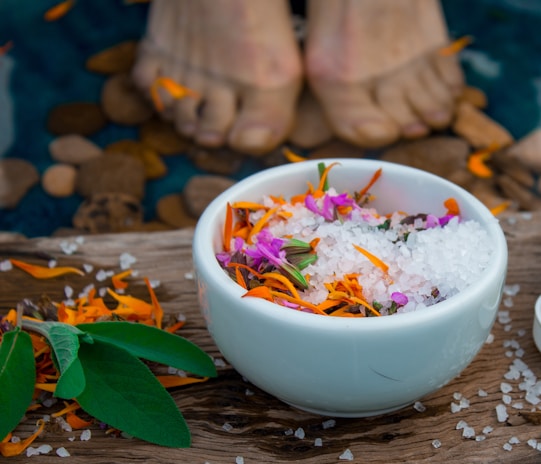 A white bowl filled with coarse salt and colorful flower petals, placed on a wooden surface. Leaves and scattered petals adorn the area around the bowl. Partially visible human feet and smooth stones are in the background, submerged in water.