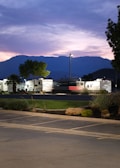A parking lot with recreational vehicles and a few trees is set against a backdrop of mountains during twilight. The sky is filled with shades of purple and pink, and a single streetlamp is lit, casting a warm glow over the area.