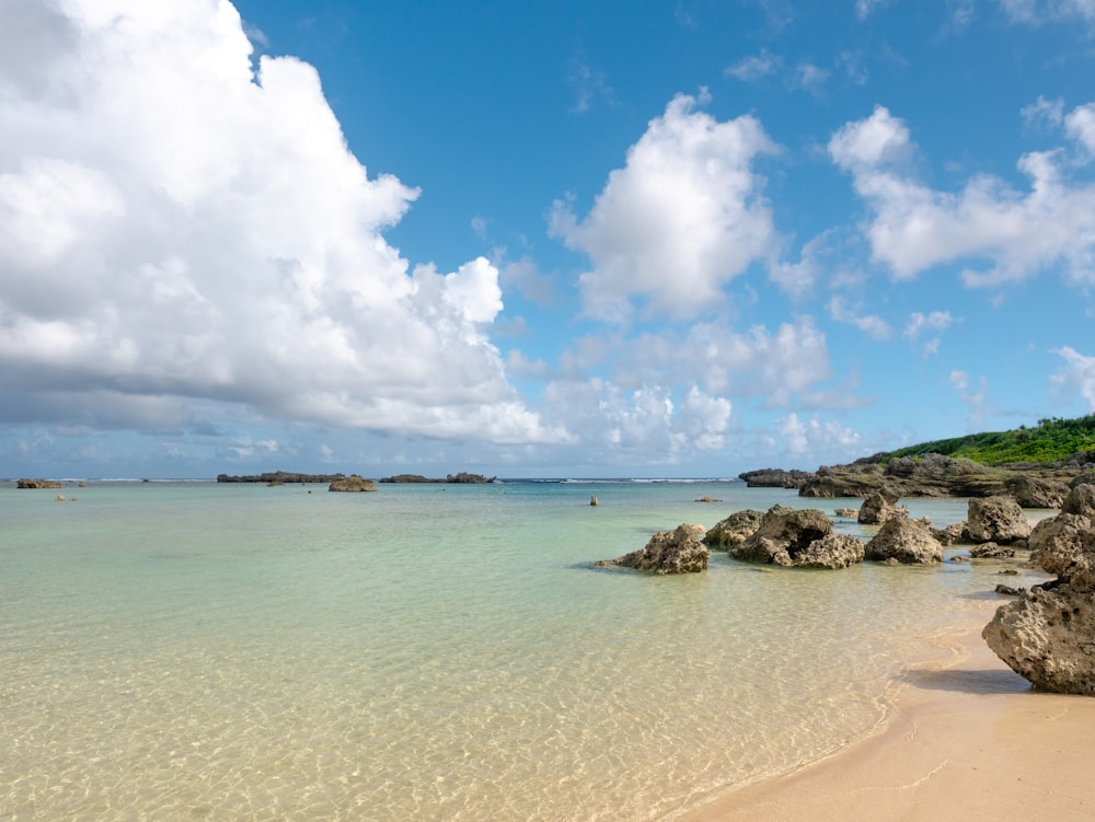 a beach with rocks and clear water under a cloudy blue sky