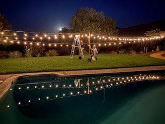 A nighttime scene featuring a backyard with decorative string lights hanging over a well-manicured lawn. Two people, one standing on a ladder and the other holding it steady, are adjusting the lights. A swimming pool in the foreground reflects the warm glow of the lights. The moon is partially visible, surrounded by trees and shrubs in the background.