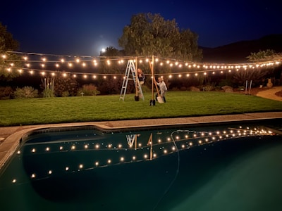 A nighttime scene featuring a backyard with decorative string lights hanging over a well-manicured lawn. Two people, one standing on a ladder and the other holding it steady, are adjusting the lights. A swimming pool in the foreground reflects the warm glow of the lights. The moon is partially visible, surrounded by trees and shrubs in the background.