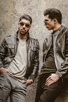 Two men wearing leather jackets and sunglasses are leaning against a rough-textured wall. One man stands with a serious expression, hands in pockets, while the other looks toward him, posing stylishly.