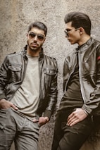 Two men wearing leather jackets and sunglasses are leaning against a rough-textured wall. One man stands with a serious expression, hands in pockets, while the other looks toward him, posing stylishly.
