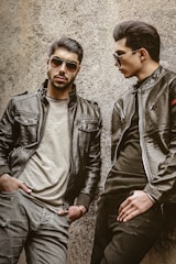 Two men wearing leather jackets and sunglasses are leaning against a rough-textured wall. One man stands with a serious expression, hands in pockets, while the other looks toward him, posing stylishly.