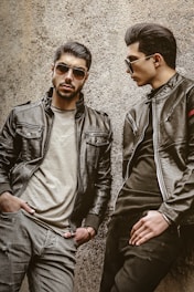 Two men wearing leather jackets and sunglasses are leaning against a rough-textured wall. One man stands with a serious expression, hands in pockets, while the other looks toward him, posing stylishly.