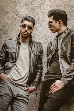 Two men wearing leather jackets and sunglasses are leaning against a rough-textured wall. One man stands with a serious expression, hands in pockets, while the other looks toward him, posing stylishly.