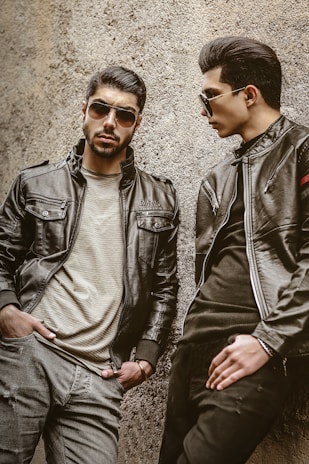 Two men wearing leather jackets and sunglasses are leaning against a rough-textured wall. One man stands with a serious expression, hands in pockets, while the other looks toward him, posing stylishly.