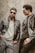 Two men wearing leather jackets and sunglasses are leaning against a rough-textured wall. One man stands with a serious expression, hands in pockets, while the other looks toward him, posing stylishly.