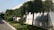 Neatly trimmed trees line the edge of a building with white tiled walls. The grass and pavement form a clean path around the hedges and trees, suggesting a maintained landscape.