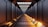 A long, futuristic corridor extends into the distance, flanked by sleek, dark walls. The floor and walls are illuminated by a series of glowing orange lights, creating a warm contrast against the cool metallic surfaces. The ceiling is patterned with a grid of recessed lighting, adding to the modern, high-tech aesthetic.