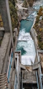 A hydroelectric dam channels water through a steep concrete structure with teal handrails. The river below is surrounded by rocky terrain and dense trees. The forceful flow of water creates foamy rapids as it descends. In the background, a bridge crosses the river amidst the rugged landscape.