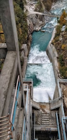 A hydroelectric dam channels water through a steep concrete structure with teal handrails. The river below is surrounded by rocky terrain and dense trees. The forceful flow of water creates foamy rapids as it descends. In the background, a bridge crosses the river amidst the rugged landscape.