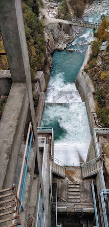 A hydroelectric dam channels water through a steep concrete structure with teal handrails. The river below is surrounded by rocky terrain and dense trees. The forceful flow of water creates foamy rapids as it descends. In the background, a bridge crosses the river amidst the rugged landscape.