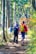 A group of people is hiking through a lush forest path surrounded by tall trees and dense undergrowth. The sunlight filters through the leaves, creating a pattern of light and shadow on the ground. The hikers are dressed in colorful outdoor attire, each carrying a backpack.