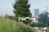 A white drone is hovering in the foreground against a backdrop of greenery and an urban skyline in the distance. The scene includes tall grass and a blurred background featuring buildings and a person riding a bike.
