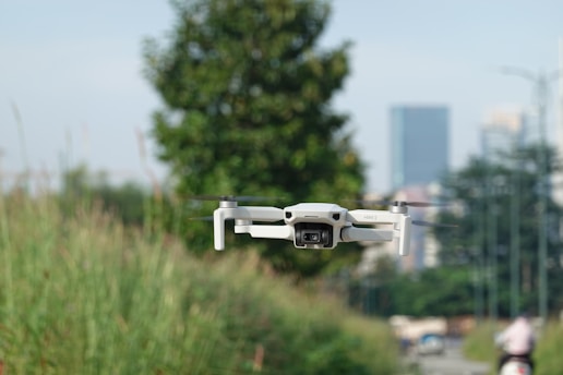 A white drone is hovering in the foreground against a backdrop of greenery and an urban skyline in the distance. The scene includes tall grass and a blurred background featuring buildings and a person riding a bike.