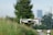A white drone is hovering in the foreground against a backdrop of greenery and an urban skyline in the distance. The scene includes tall grass and a blurred background featuring buildings and a person riding a bike.