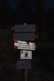 A wooden post displays several directional signs with information for hikers. The signs list destinations such as Villa Welsperg, Rif. Pradidali, Malga Canali, Rud. M. Pradidali, and Rif. Canali-Treviso, along with the corresponding distances. The bottom sign indicates a hiking trail for experienced hikers. The background is dark, with hints of trees and foliage.