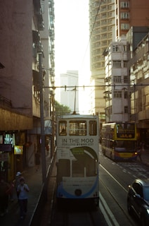A busy urban street scene with a white tram in the center, bearing an advertisement for 'In the Moo for Love 2021'. The tram is flanked by tall residential buildings bathed in warm sunlight. A double-decker bus is visible on the side, alongside pedestrians walking on the sidewalk.