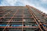 A perspective view looking up at a building facade with extensive scaffolding. The scaffolding consists of metal beams and platforms, creating a grid-like pattern. The sky in the background is partly cloudy with a blue hue.