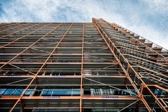 A perspective view looking up at a building facade with extensive scaffolding. The scaffolding consists of metal beams and platforms, creating a grid-like pattern. The sky in the background is partly cloudy with a blue hue.