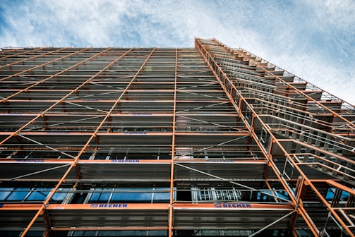 A perspective view looking up at a building facade with extensive scaffolding. The scaffolding consists of metal beams and platforms, creating a grid-like pattern. The sky in the background is partly cloudy with a blue hue.