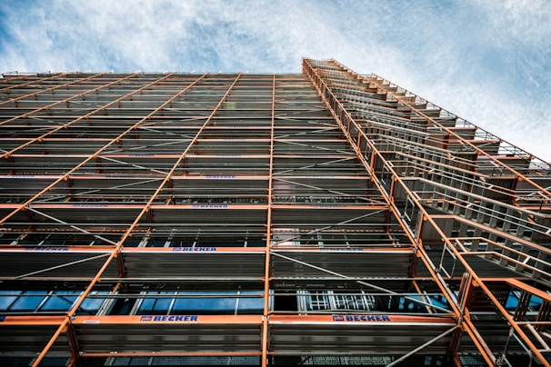 A perspective view looking up at a building facade with extensive scaffolding. The scaffolding consists of metal beams and platforms, creating a grid-like pattern. The sky in the background is partly cloudy with a blue hue.