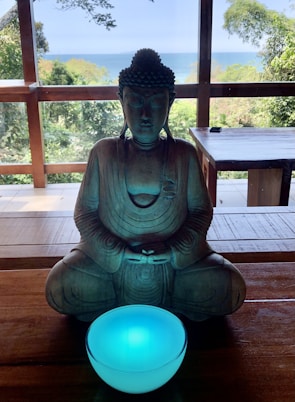 A stone statue of a meditative figure in a seated position, holding a glowing blue bowl. The statue is placed on a wooden table with a backdrop of lush greenery and a distant view of the ocean under a clear sky.