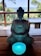 A stone statue of a meditative figure in a seated position, holding a glowing blue bowl. The statue is placed on a wooden table with a backdrop of lush greenery and a distant view of the ocean under a clear sky.