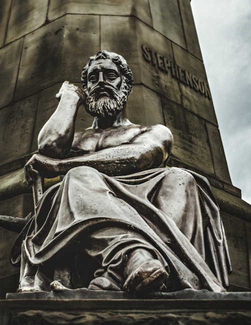 A large statue of a contemplative bearded man, sitting with his chin resting on his hand. The sculpture is detailed with flowing robes and an expression of deep thought. The backdrop features a tall stone pedestal with a name inscribed on it.