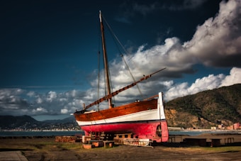 A large wooden sailboat is positioned on land, supported by wooden blocks. The boat's hull is painted in red and white, with a natural wooden finish on the deck and mast. In the background, a picturesque landscape features distant mountains, a calm sea, and a small coastal town under a dramatic sky filled with fluffy white clouds.