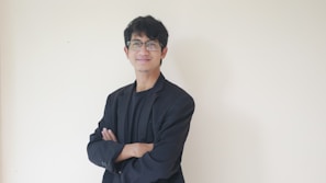A person stands confidently against a plain light-colored background, wearing glasses and a black suit with a black shirt. Their arms are crossed, and they are smiling slightly, projecting a calm and approachable demeanor.