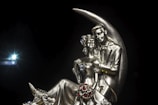 A metallic sculpture of a couple seated together, intricately designed with detailed features and decorative elements. The figures are positioned against a crescent moon backdrop, with elegantly sculpted attire adorned with small jewel-like details. The background is a solid black, enhancing the focus on the sculpture.